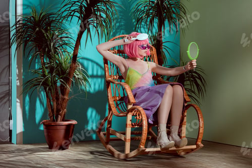 Preview: attractive girl in pink wig combing hair and looking at mirror while sitting in rocking chair