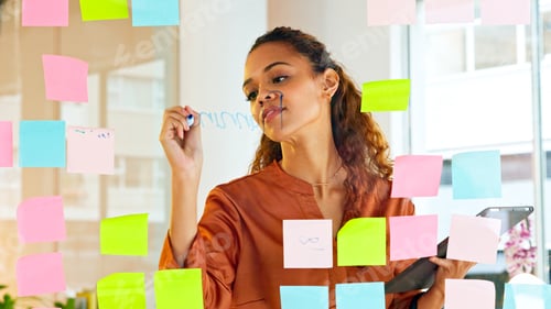 Preview: Focused female designer planning ideas on a glass wall with colorful sticky notes inside a modern a