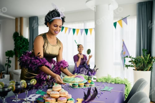Preview: Happy black woman serving King cake at dining table during Mardi Gras celebration party at home.
