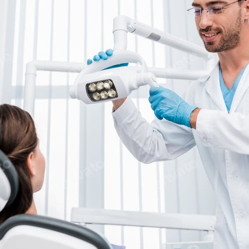 Preview: selective focus of handsome dentist in latex gloves touching medical lamp near patient