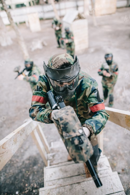 Preview: high angle view of paintball player aiming by marker gun and running on staircase while his team
