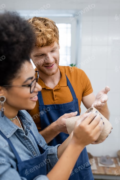 Preview: happy redhead man pointing with hand at clay cup during date with african american girlfriend in