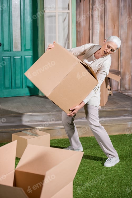 Preview: Senior woman standing on grass and holding big cardboard box while moving into new house