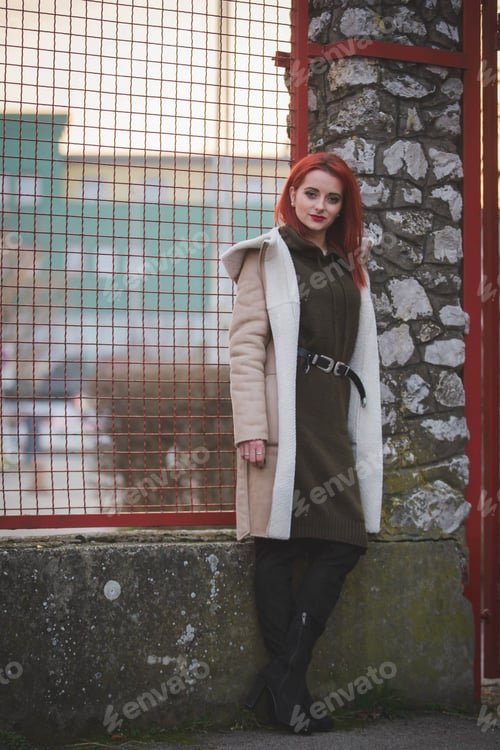 Preview: Red-haired female in a jacket with a hood posing in front of a grid fence