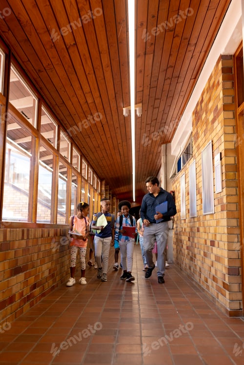Preview: Diverse male teacher and children walking together in corridor at elementary school, copy space