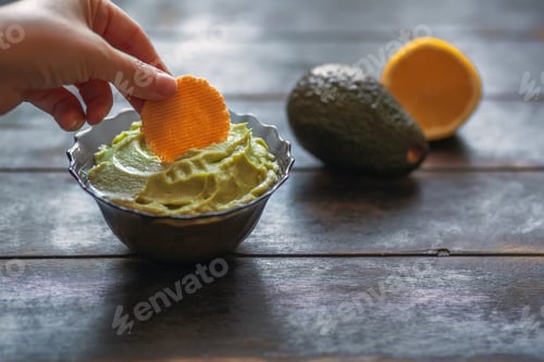 Preview: Woman dips a cracker in the mexican sauce guacamole in bowl, avocado and lemon on dark background