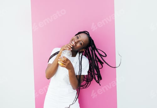 Preview: Cheerful woman with braids drinking a juice and looking at camera while standing at pink wall