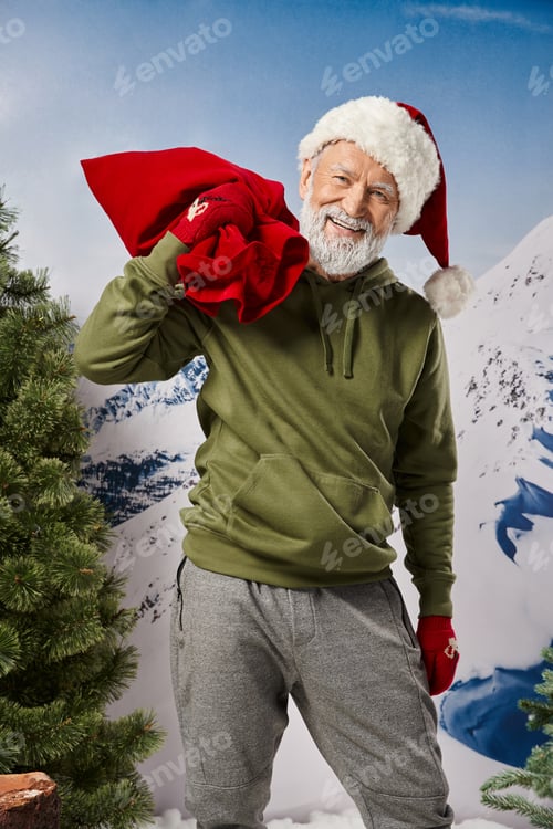 Preview: joyful Santa with christmassy hat and present bag smiling cheerfully at camera, winter concept