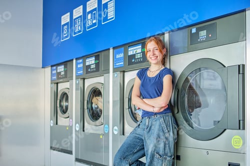 Preview: Smiling young redhead woman standing in a public laundromat looking at camera