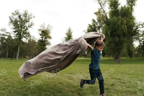Preview: Cheerful boy running with a flying blanket in the park
