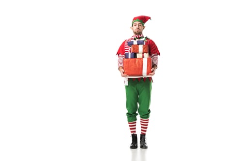 Preview: man in christmas elf costume looking at camera and carrying pile of presents isolated on white