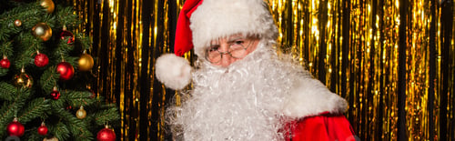 Preview: Bearded santa claus looking at camera near christmas tree and tinsel, banner
