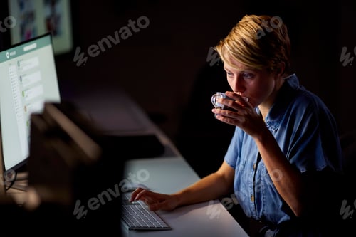 Preview: Businesswoman Working Late In Office With Face Illuminated By Computer Screen Drinking Coffee