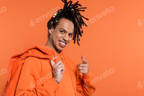 Preview: smiling multiracial man in hoodie holding denim jacket and pointing at camera isolated on coral
