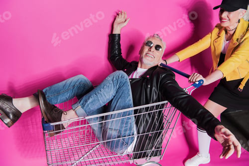 Preview: Smiling stylish senior woman pushing shopping trolley with joyful senior man having fun