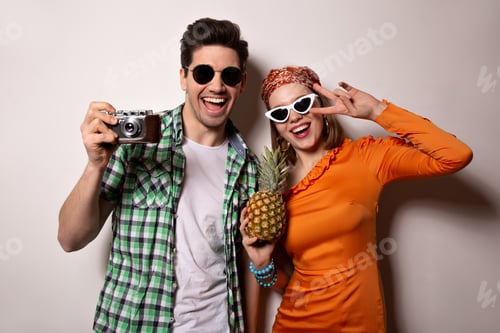 Preview: Active couple in bright summer outfits and sunglasses posing with retro camera and pineapple. Girl