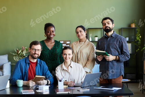 Preview: Team of young successful designers gathered by workplace looking at camera