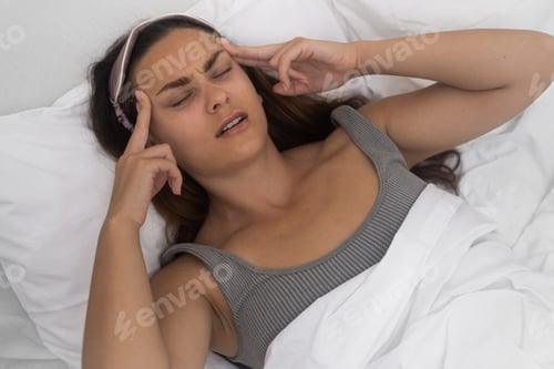 Preview: A lady in grimaces, temples aching, hands cradling her head, battling a headache on a bright bed.