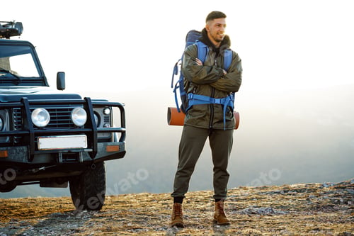 Preview: Portrait of a young traveler man in hiking equipment standing near his off-road car