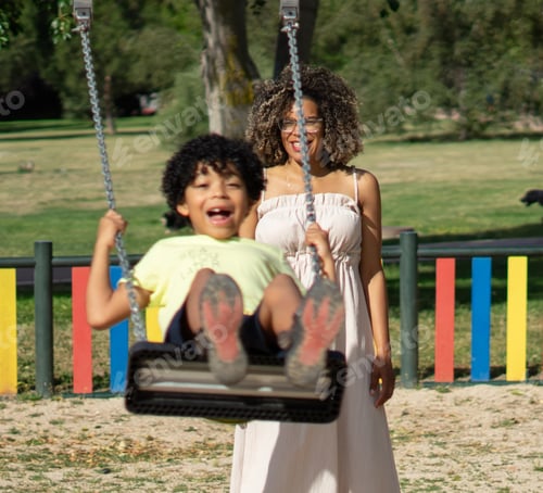 Preview: Latin mother with afro hair swinging her son on a playground swing