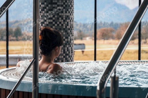 Preview: Rear view of young woman relaxing in a jacuzzi in spa inside a hotel resort with beautiful view.