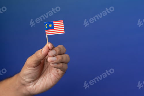 Preview: A hand holds a small miniature flag of Malaysia, Jalur Gemilang against a solid blue background.