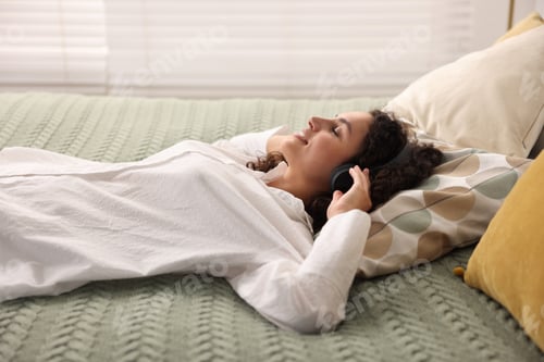 Preview: Young woman with headphones listening to music on bed at home