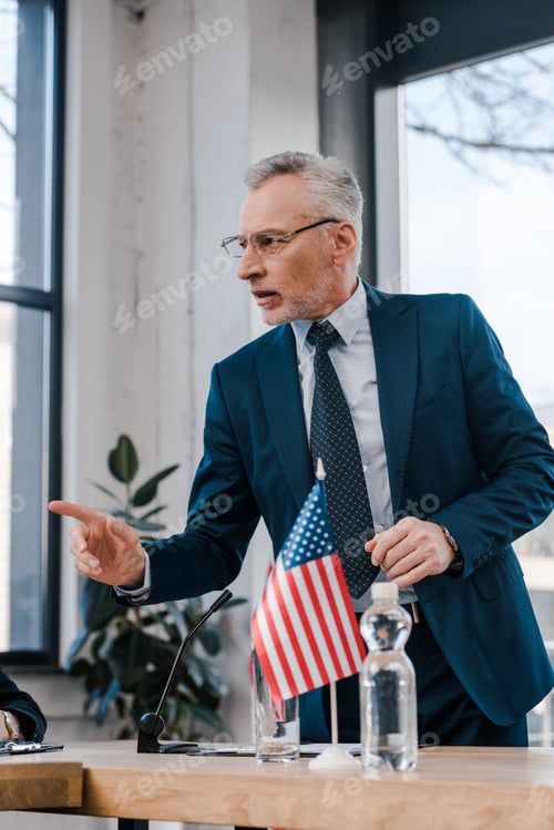 Preview: selective focus of bearded diplomat in eyeglasses pointing with finger while talking near american