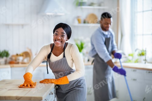 Preview: Portrait of happy black lady in apron and rubber gloves wiping kitchen table, her boyfriend washing