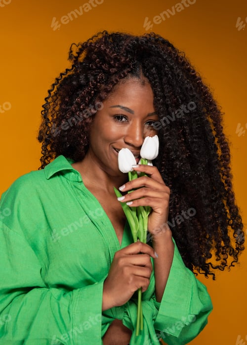 Preview: Beautiful African american woman in green shirt holds white tulips and smiles on yellow background