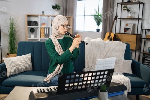 Preview: Young muslim woman in hijab plays flute on bright living room at home.