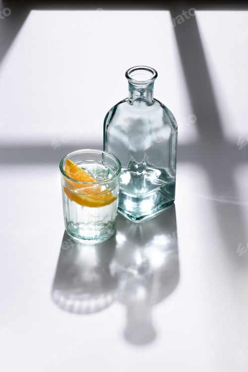 Preview: close up view of glass with lemon piece and bottle with water on white surface