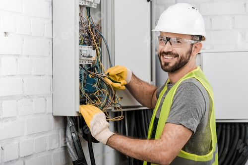 Preview: smiling handsome electrician repairing electrical box with pliers in corridor and looking at camera