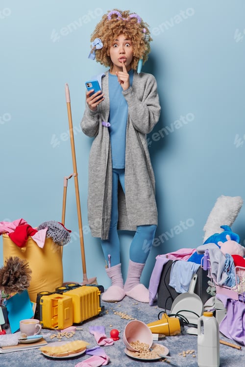 Preview: Vertical shot of mysterious housewife makes silence gesture dressed in domestic clothes holds mobile