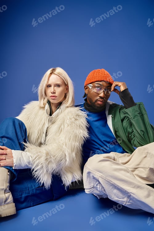 Preview: young african american man and blonde woman in winter attire sitting together on blue backdrop