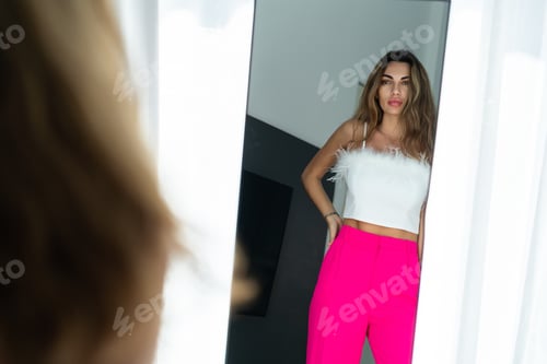 Preview: Stylish fit tanned beautiful woman in fashion pink pants and top posing in mirror at home bedroom