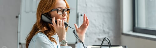 Preview: Redhead businesswoman gesturing while talking on landline telephone with blurred flipchart on