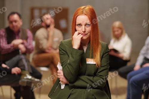 Preview: Unhappy and crying middle aged woman in casualwear standing against patients of mental support group