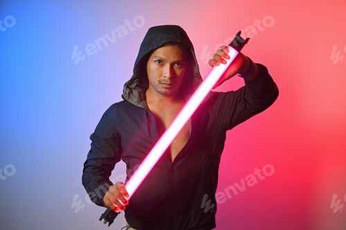 Preview: Portrait of handsome man in hoodie holding LED neon light stick against red and blue background.