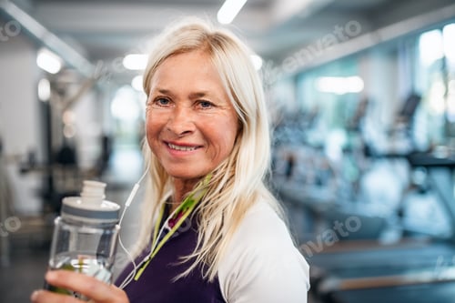 Preview: A senior woman with earphones and water bottle standing in gym