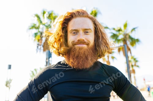 Preview: handsome redhaireb man with long beard wear black sporty suit and warming up in sunny beach