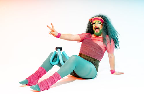 Preview: Smiling Drag Queen with long hair sitting on ground exercicing legs and doing victory sign with hand