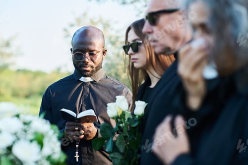 Preview: Focus on serious priest in black attire looking in open Bible at funeral service