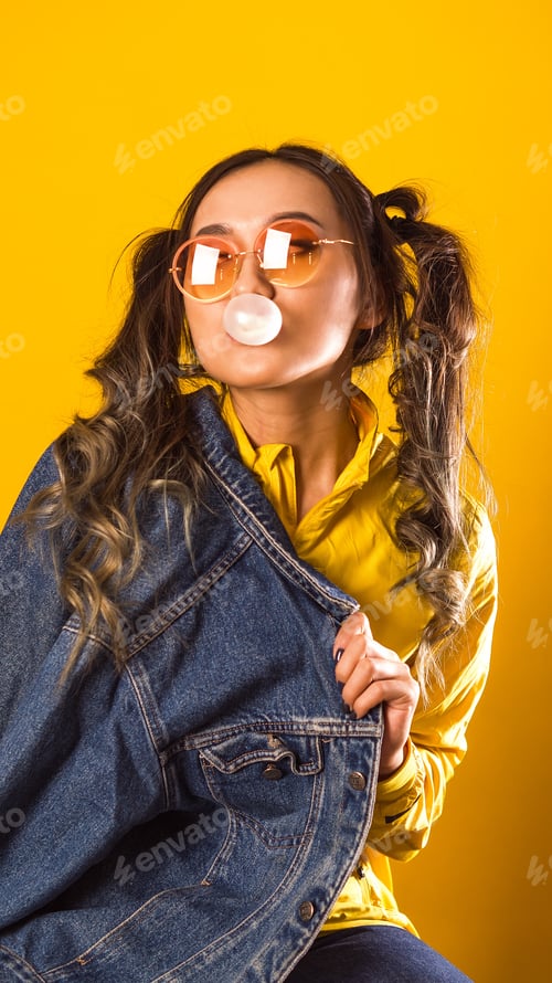 Preview: Happy woman blowing a soap bubble with chewing gum on a yellow background.