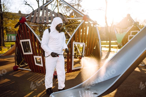 Preview: Man wearing protective suit disinfecting the playground in the sun with spray. Covid -19. Cleaning.