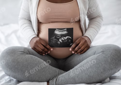Preview: Unrecognizable black pregnant lady demonstrating her baby sonography photo, sitting on bed