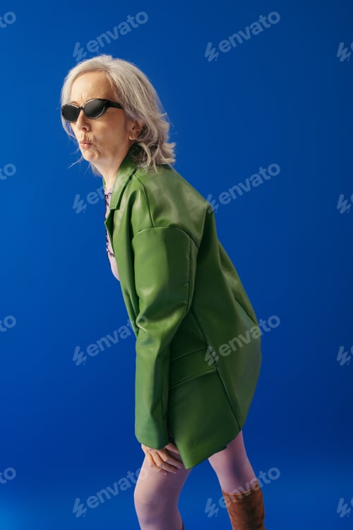 Preview: grey haired woman in stylish sunglasses and green leather jacket pouting lips isolated on blue