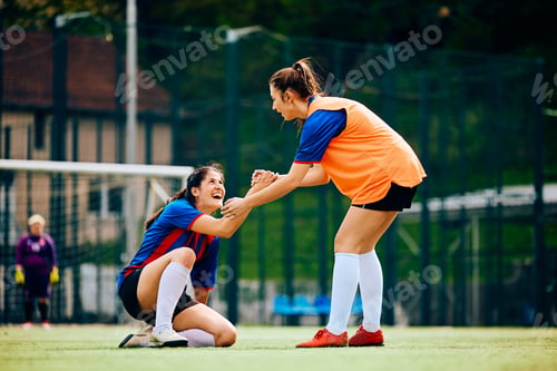 Preview: Happy female player helping her teammate to get up on soccer pitch.