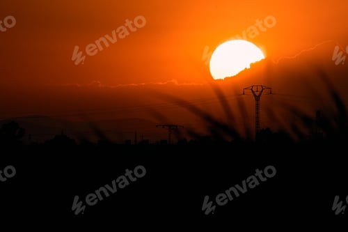 Preview: impressive sunset with the sun hiding behind the clouds over some plants and high voltage towers