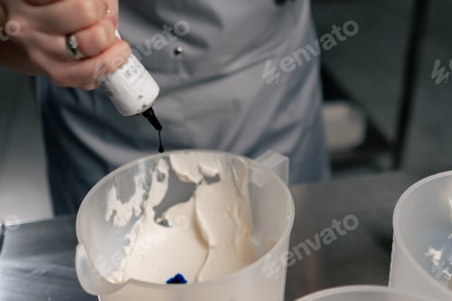 Preview: close-up of a female baker in a professional kitchen adding blue coloring to white cream